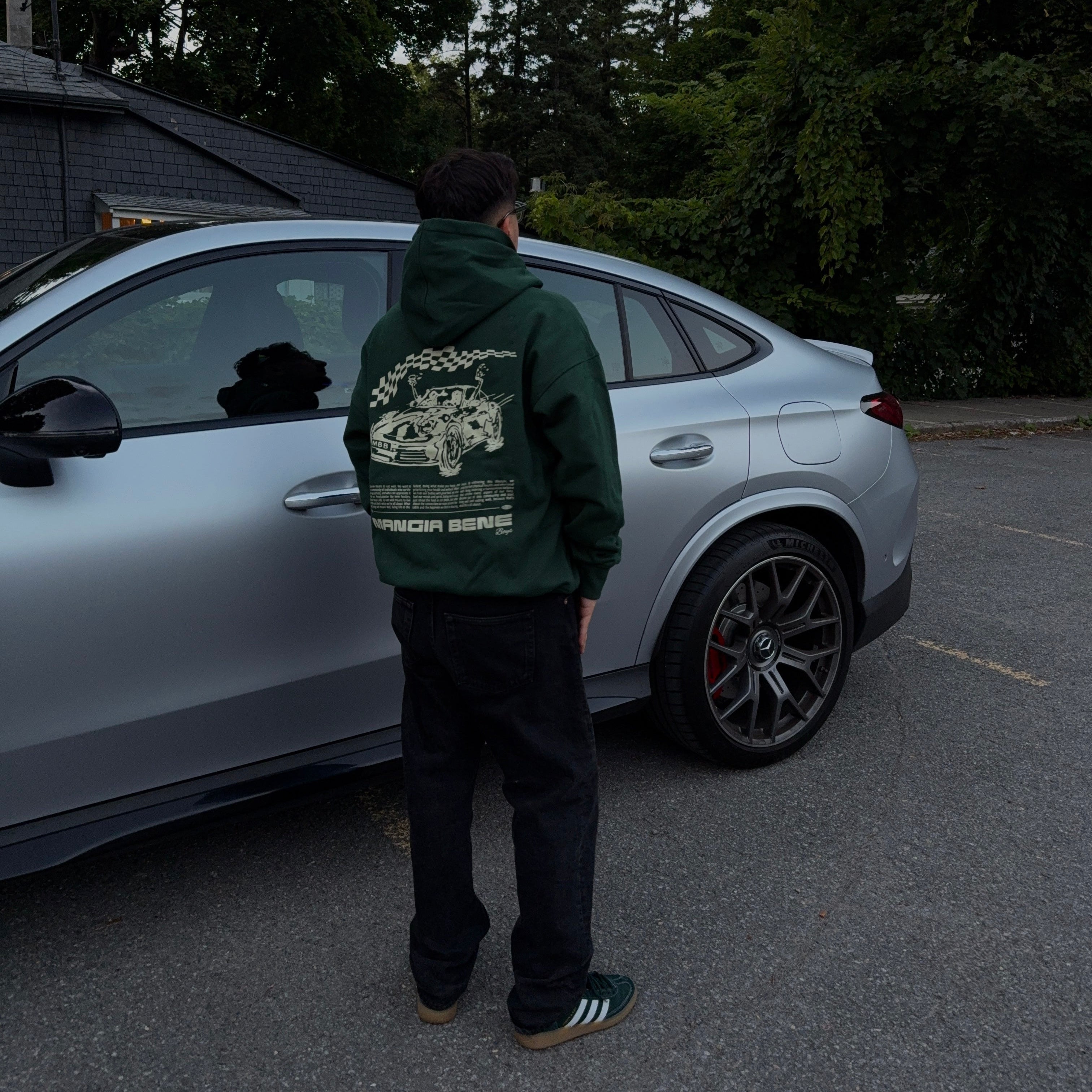 Person wearing a green hoodie with a car design, standing next to a silver car in a parking lot with trees in the background.
