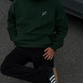 Person wearing a dark green hoodie and black pants sitting on a dark surface.