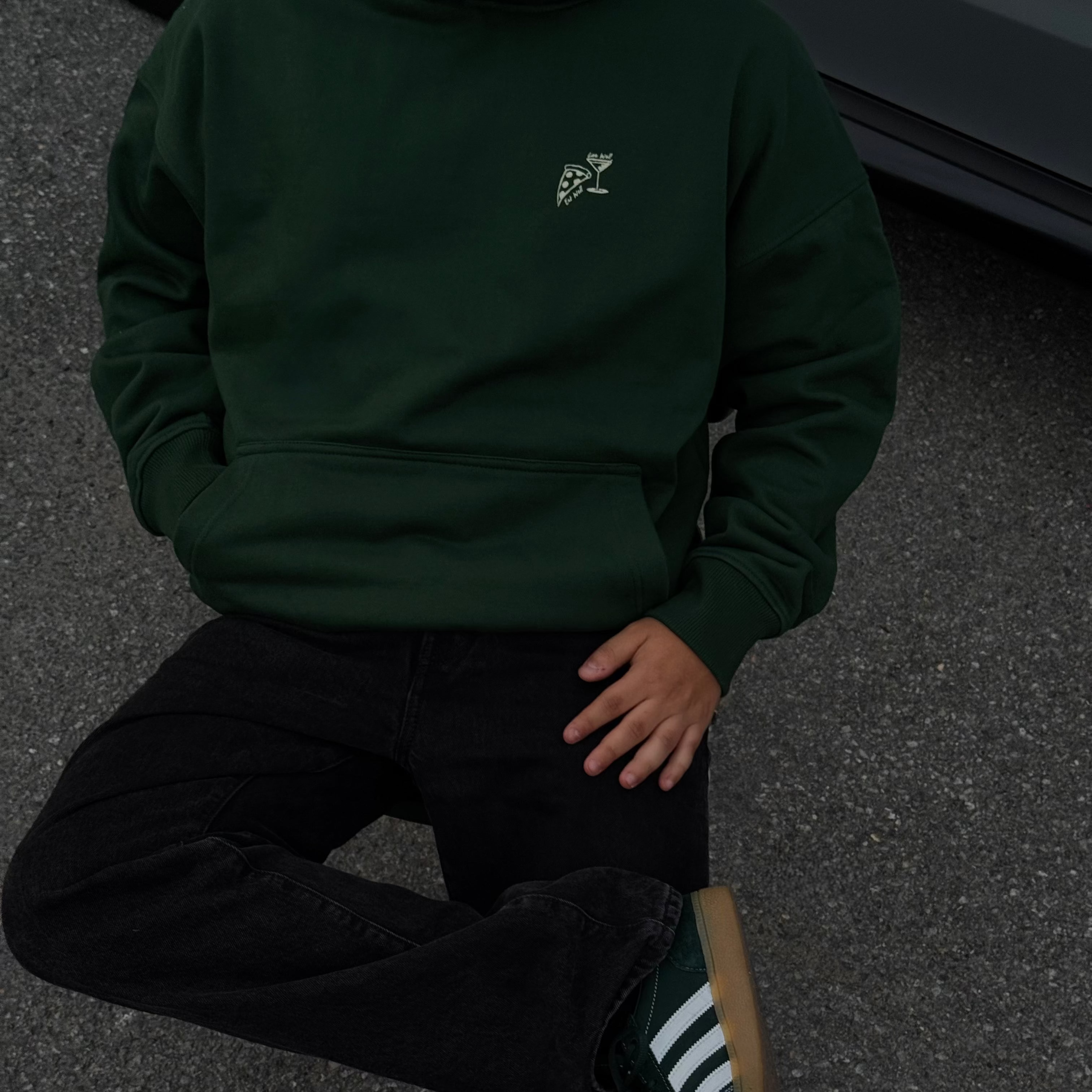 Person wearing a dark green hoodie and black pants sitting on a dark surface.